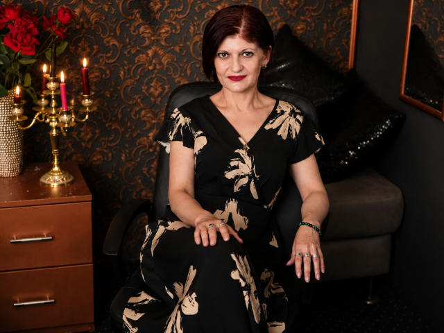 MichelleNoir con capelli rossi, vestito floreale nero, seduta su una sedia, sfondo nero, comò di legno con portacandele, rose rosse, cuscino nero, carta da parati nera a motivi, sorridente, rossetto rosso..