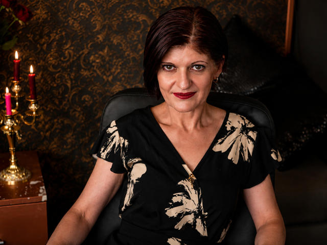 MichelleNoir, uma mulher de meia-idade com cabelo castanho curto, batom vermelho, vestido floral preto, sentada em uma cadeira, olhando para a câmera, um candelabro dourado com velas vermelhas à esquerda..