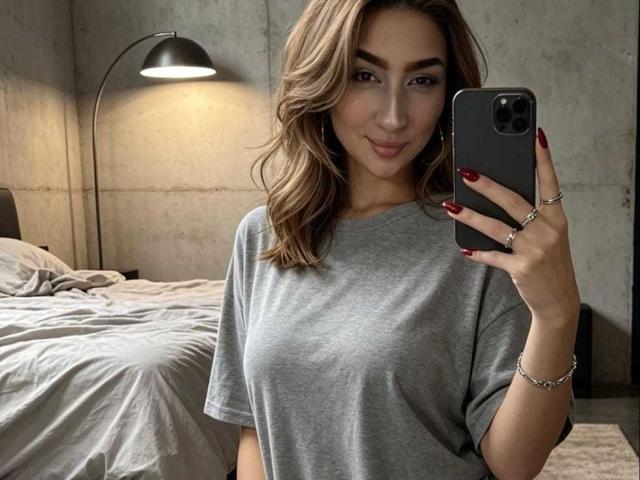 Abby, una mujer con cabello castaño ondulado, uñas rojas, camiseta gris, sosteniendo un teléfono, sonriendo, con senos de tamaño medio, un anillo en el dedo, una cama al fondo, una pared de concreto, una lámpara negra, luz suave..