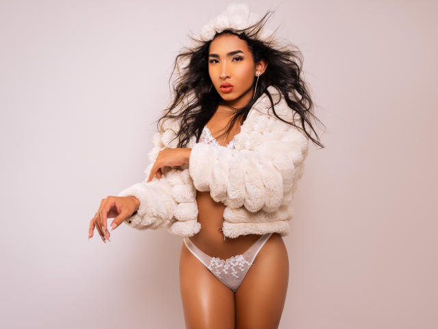 Gaby Santos, com longos cabelos pretos, vestindo uma jaqueta branca e fofa, calcinhas de renda brancas e uma faixa de cabelo branca, exibindo seus seios e vulva, com um olhar confiante e sexy..