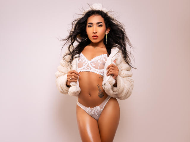 Gaby Santos, con cabello negro largo, vestida con lencería blanca de encaje, una chaqueta beige esponjosa y aretes de aro dorados, mostrando un tatuaje en la cadera izquierda, está de pie con las manos sobre la chaqueta..