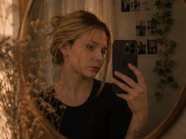 LeeMako met blond haar in een rommelige knot, die een spiegelzelfportret maakt, een zwarte telefoon vasthoudend, een zwarte top dragend, met een serieuze blik, met gedroogde bloemen op de voorgrond..