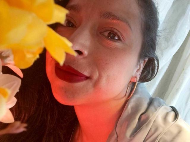 StangelarRivera's face with red lipstick, dark hair, hoop earrings, and a white hoodie, holding yellow flowers near her nose, smiling slightly, with a red light on her cheek.