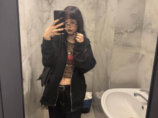 Haydee Doncaster mit dunkelbraunen Haaren, Brille und schwarzem Lippenstift, trägt eine schwarze Jacke, ein braunes Oberteil und eine schwarze Hose und macht ein Selfie vor einem Spiegel im Badezimmer..
