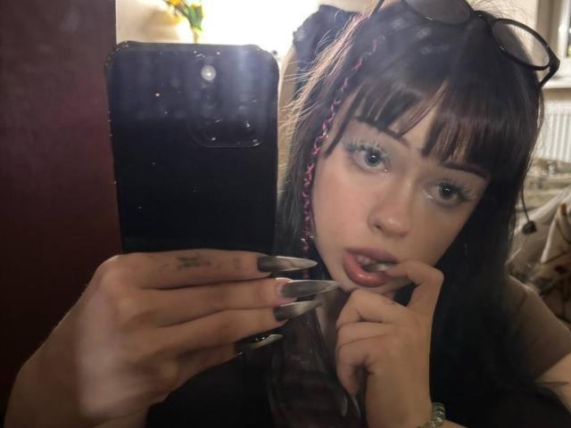 Haydee Doncaster, con capelli castano scuro, frangia e trecce rosa, che si guarda in uno specchio con unghie lunghe, mentre si morde un dito e tiene in mano un telefono con la fotocamera ben visibile..