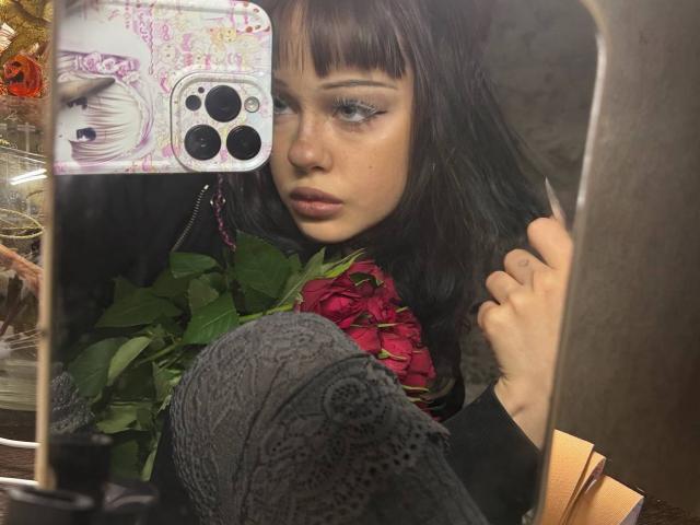 Haydee Doncaster mit dunkelbraunen Haaren und Pony, trägt ein schwarzes Oberteil, graue gestrickte Beinwärmer und hält ein Handy mit einer pink-weißen Blumenhülle. Sie blickt in einen Spiegel..