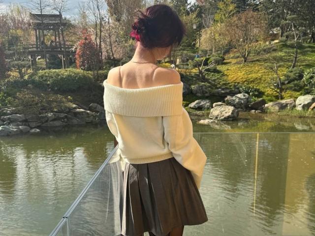 HaydeeDoncasters blanker Rücken zeigt einen sexy, schulterfreien weißen Pullover, einen schwarzen Rock und einen roten Dutt. Sie steht an einem Teich mit Steinen und Grünpflanzen. Ihr Hintern ist teilweise sichtbar..