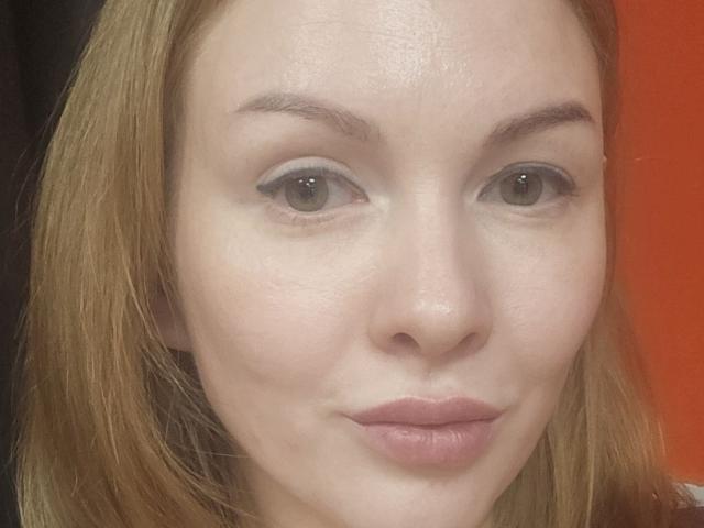 Het gezicht van KatyBrie, met lichte huid, groene ogen en blond haar, toont een lichte glimlach en roze lippen, en ze kijkt recht in de camera. Haar achterkant is niet zichtbaar..