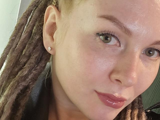 KatyBrie's face with brown dreadlocks, green eyes, subtle makeup, pink lipstick, small diamond earrings, and a slight smile. Her skin is fair and slightly shiny.