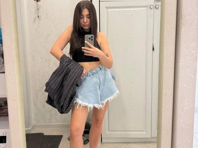 InesGueningsman with long brown hair, black crop top, and frayed blue denim skirt, holding striped jacket, taking mirror selfie, showing her ass and small tits.
