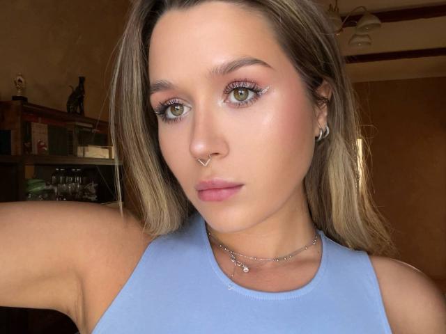 Viktoriyya, mit grünen Augen, trägt ein blaues Tanktop, hat eine Nasenpiercing und kleine Ohrringe, hat hellbraunes Haar und einen dezent rosafarbenen Lippenstift und blickt in die Kamera..