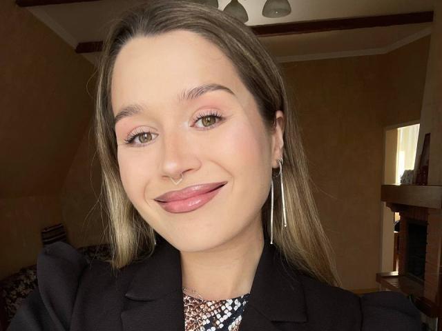 Viktoriyya, com cabelo castanho claro, sombra rosa, olhos verdes, piercing no nariz, lábios rosados brilhantes, jaqueta preta e brincos longos, sorrindo para a câmera..