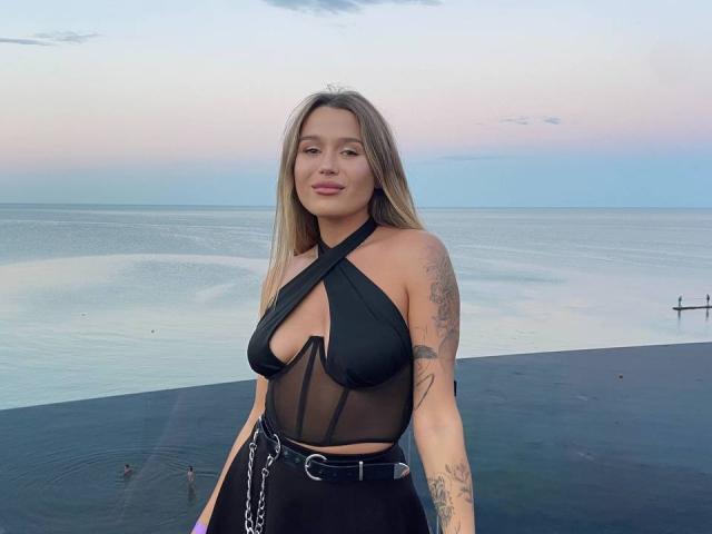 Viktoriyya, con i capelli biondi, un abito nero a fascia che mette in mostra il seno e dei tatuaggi, è in piedi accanto a una piscina nera a forma di infinity al tramonto, indossando una cintura nera con una catena..