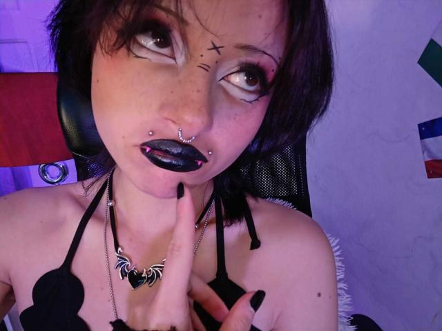 ViolettClay with black lipstick, nose ring, and lip piercing, wearing a black halter top, finger on chin, looking up with heavy makeup, black nails, and a butterfly necklace.