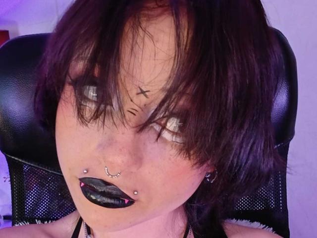 ViolettClay tem cabelos escuros e desalinhados, delineador preto, batom preto, piercing no nariz, piercing nos lábios e uma cadeira de malha preta com apoio de cabeça de couro. Pele clara, levemente avermelhada, com piercings visíveis no nariz e nos lábios..