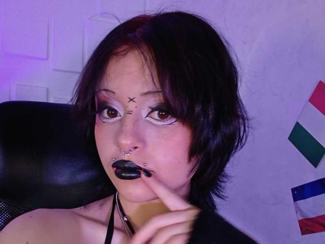 ViolettClay with black lipstick, nose piercings, and a septum piercing, holding her finger to her lips, wearing a black halter top, short dark hair, and purple eye makeup.