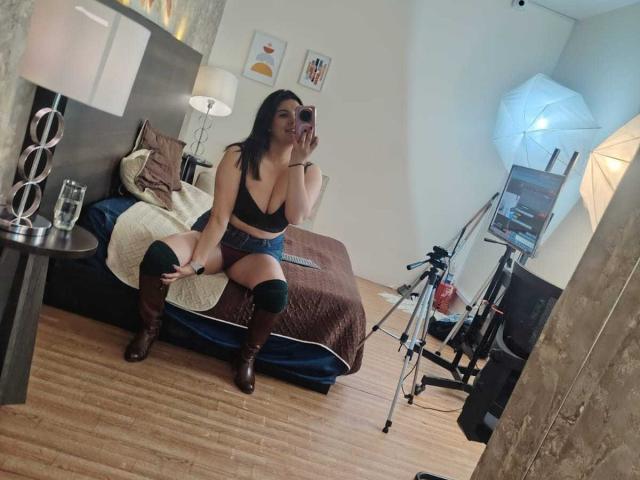 ZoeyGrey with tits in a black bra, wearing blue shorts, brown boots, and black thigh-highs, sitting on a bed, taking a mirror selfie, in a wooden-floored room.
