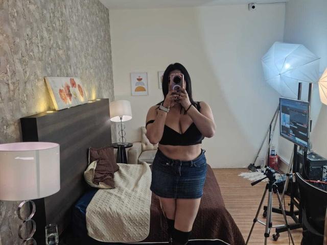 ZoeyGrey, mirror selfie, black bra, denim skirt, black boots, holding phone, covering face, small black tattoo on upper arm, bedroom with bed, lamps, camera, white umbrella.