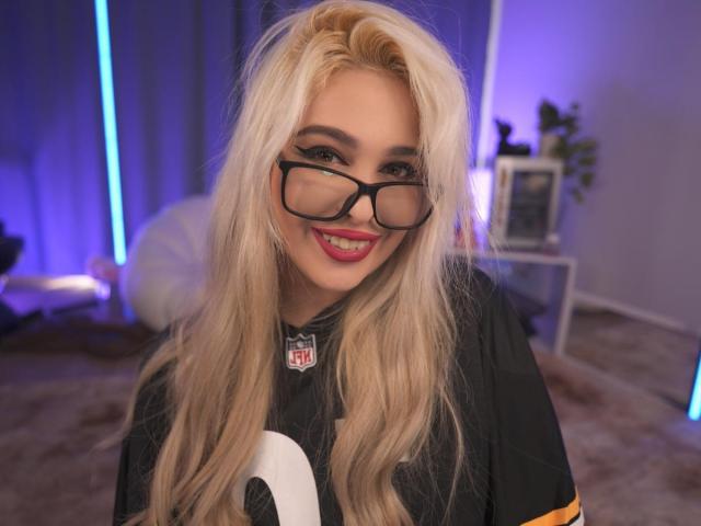 BiiaKristal, con cabello rubio, gafas, lápiz labial rojo y una camiseta deportiva negra, sonriendo, mostrando ligeramente sus senos, en una habitación con iluminación azul..