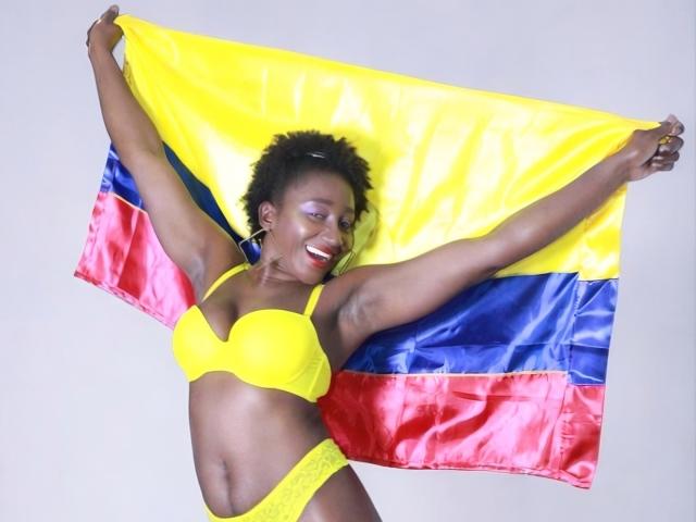 NahomyHawk met donkere huid, een gele bh, een gele onderbroek, die de Colombiaanse vlag vasthoudt, lachend, met armen omhoog, kort krullend haar, met oorringen, staand voor een effen achtergrond..
