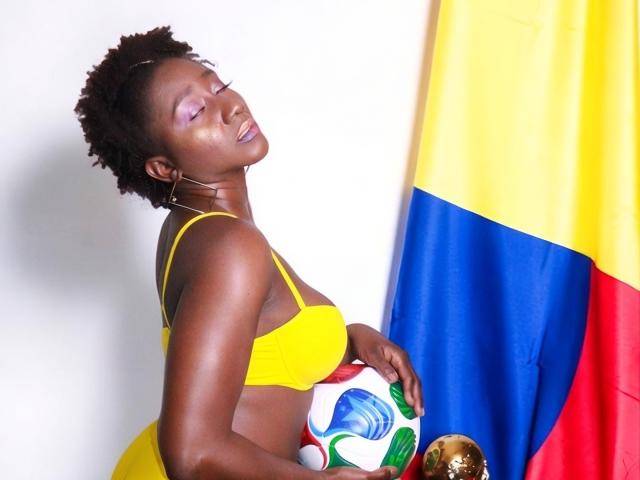 NahomyHawk, com pele escura, cabelo curto e cacheado, usando um sutiã amarelo, segurando uma bola de futebol, olhos fechados, vestido amarelo, brincos grandes de argola dourados, uma bandeira amarela e azul à direita..