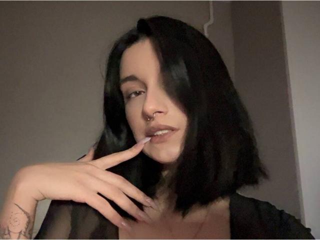 MackenzieCostantino with black shoulder-length hair, nose ring, and tattoo on finger, holding cigarette, looking at camera, wearing sheer black top, pale skin, slightly parted lips.