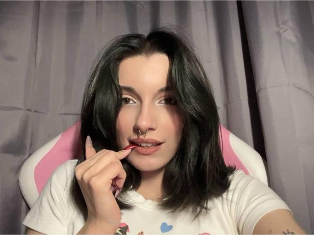 MackenzieCostantino with dark, wavy hair, nose ring, and pink lip, biting her red fingernail, wearing a white shirt, sitting in a pink and white chair.