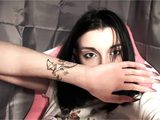 MackenzieCostantino with black hair, pale skin, and brown eyes, covering her mouth with a tattooed arm. She's wearing a pink and white chair, showing a floral shirt.