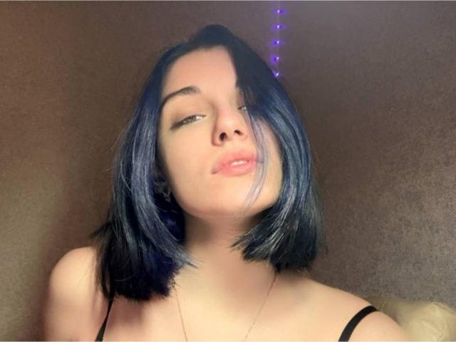 MackenzieCostantino with black shoulder-length hair, half-closed eyes, and pouty lips, wearing a black strap top, with subtle purple lights above her head.