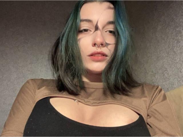 MackenzieCostantino has teal-black hair, a nose ring, and wears a brown top with a cutout, showing her tits. She's looking slightly down with a neutral expression.
