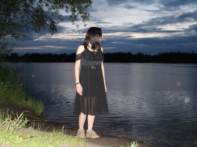 MackenzieCostantino stands by a lake at dusk, wearing a black off-shoulder dress and beige shoes, with dark hair and a black choker, looking down.