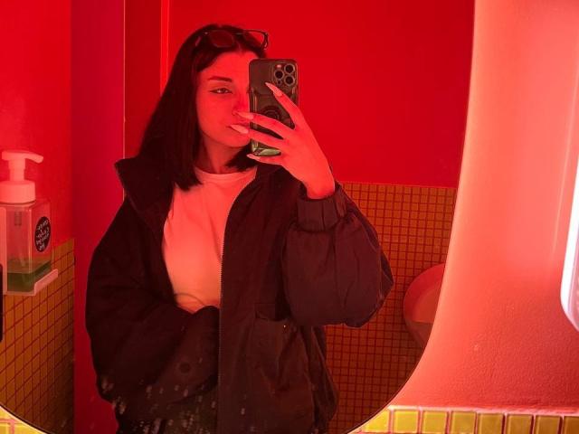 MackenzieCostantino with red-tinted mirror selfie, black jacket, white shirt, holding phone, medium-length dark hair, subtle smile, soap dispenser on left, yellow-tiled wall, mirror reflection.