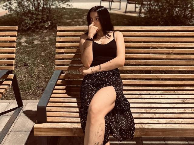 MackenzieCostantino sits on a wooden bench with a high black skirt showing her thigh. She's wearing a black tank top, covering her mouth with her hand, looking down.
