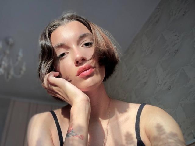 MackenzieCostantino has short brown hair, light skin, and a tattoo on her right wrist. She's wearing a black top, touching her chin, with a subtle, sexy smile.