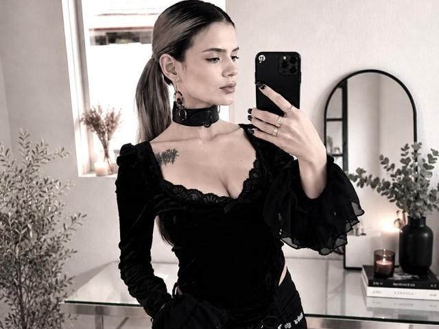 ErgoProxy vestindo um vestido de veludo preto, com mangas bufantes, um colar preto e um colar, tirando uma selfie no espelho, mostrando seios pequenos e uma tatuagem no lado direito do peito, em um quarto moderno..