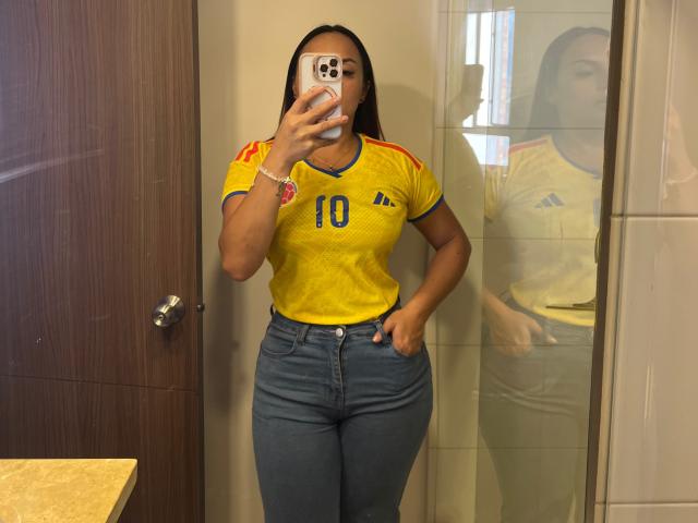 GalaMalkova with medium brown skin, in yellow soccer shirt with "10" and red stripes, blue jeans, hand in pocket, mirror selfie, reflection, phone in front, brown door.
