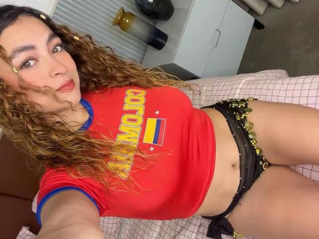 Arianna Blake mit lockigem, braunem Haar, einem roten Hemd und schwarzen, blumigen Unterhosen, die ihre Brüste und ihren Hintern zeigen, liegt auf einem Bett mit einer rosa karierten Bettdecke..
