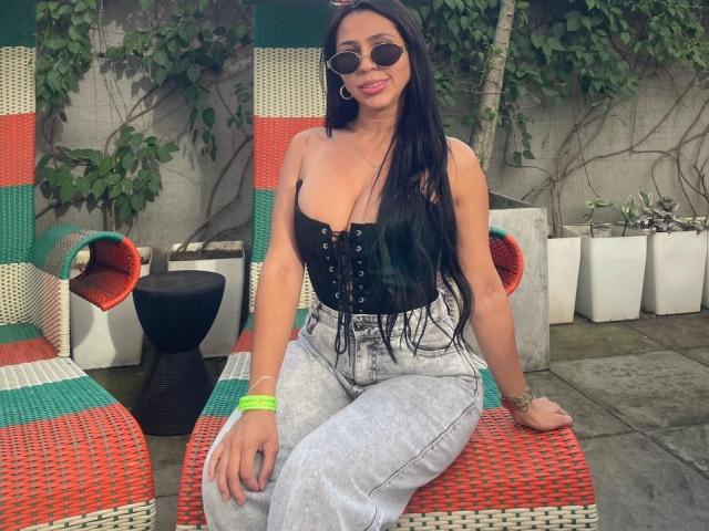 NahomyStone with long black hair, wearing round sunglasses, black lace-up bustier, and gray high-waisted jeans, sitting on colorful woven chair, showing her big boobs and cute smile.