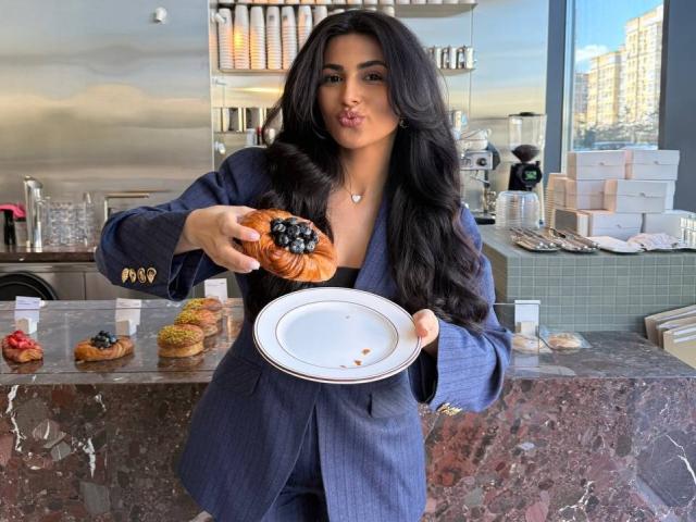 AdrianaPierce with long black hair, blue suit, puffy lips, holding a donut with blueberries, using other hand to hold a white plate, in a modern bakery.