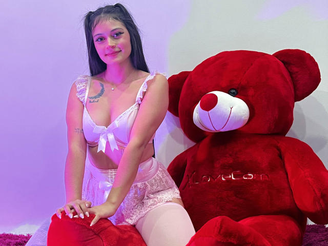 LizzaRose mit langem, schwarzem Haar, weißer Spitzenunterwäsche und rosafarbenen Strümpfen sitzt neben einem riesigen, roten Teddybären, lächelt und zeigt Tattoos an ihrer rechten Schulter und ihrem linken Arm..
