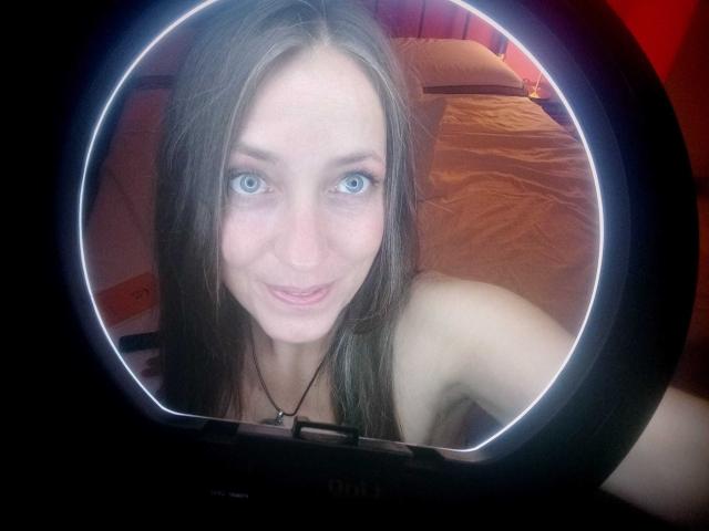 Os ombros nus de DaisyDea, seus olhos azuis e sorriso discreto são refletidos em uma luz de anel, com seu longo cabelo castanho e colar visíveis. Seus seios não são mostrados..