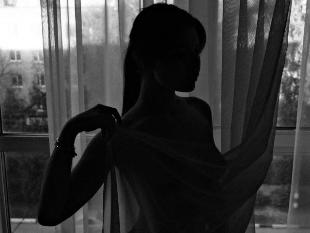 Silhouetted KizzyDepner with long hair, raising a sheer top to expose her tits, standing in front of a window with translucent curtains. Black and white photo.