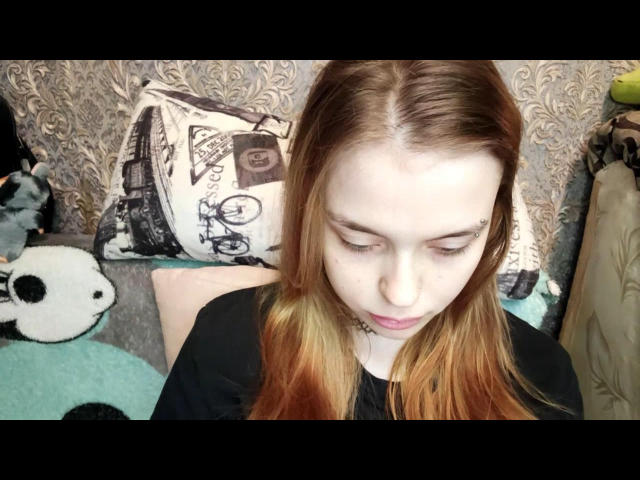 Merri Serafin hat eine blasse Haut, glattes, rotbraunes Haar und ein kleines Nasenpiercing. Sie schaut nach unten und trägt ein schwarzes Hemd mit einer Halskette um den Hals..