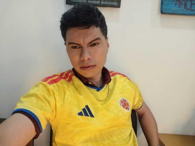 Aquiles Taylor, con i capelli neri corti, indossa una maglia da calcio gialla con bordi rossi e blu, guarda la telecamera con un leggero cipiglio, seduto su una sedia..
