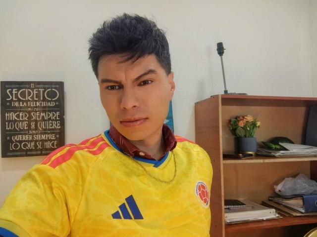 AquilesTaylor, che indossa una maglia da calcio gialla e rossa, ha i capelli neri corti e un'espressione seria, con il colletto di una camicia blu che spunta. Scaffali con una pianta sullo sfondo..