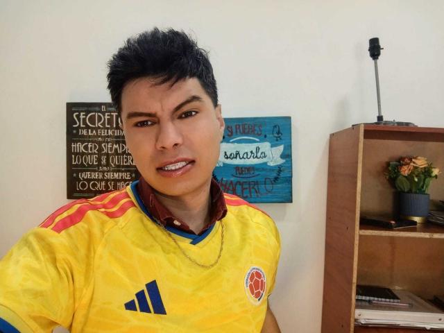 AquilesTaylor with short black hair, yellow soccer shirt with red and blue stripes, and a red emblem, smiling slightly, showing teeth, standing in front of a wooden shelf.