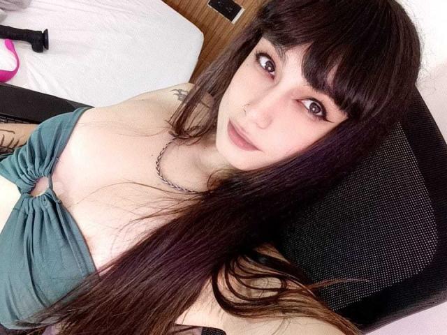 JennInk, com cabelos longos e castanhos escuros, um piercing no nariz e uma blusa verde que mostra seus seios, sentada em uma cadeira preta, olhando para a câmera com um leve sorriso..