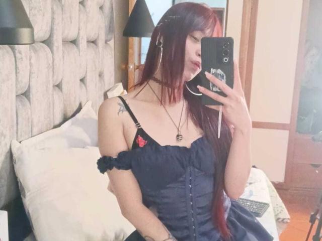 JennInk con i capelli rossi, indossa un abito nero con spalline scoperte e piccoli cuori rossi, tiene un telefono, ha tatuaggi sulla spalla, una collana, un selfie allo specchio, camera da letto, trucco leggero..