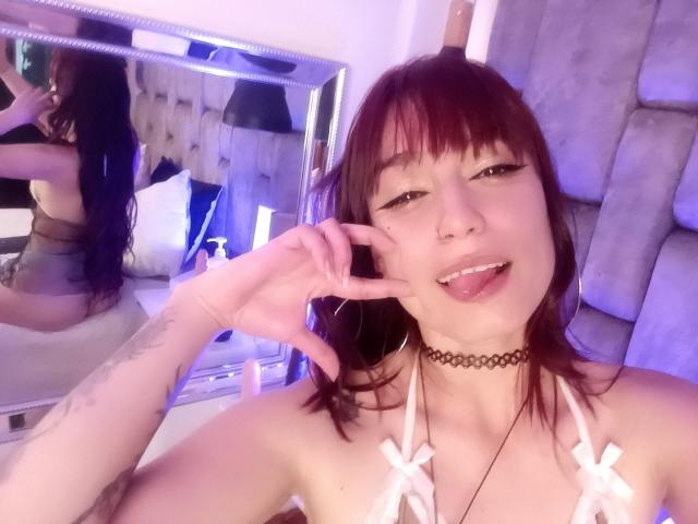 JennInk mit roten Haaren, einem schwarzen Choker, einem weißen Top mit Trägern und Schleifen, Zunge heraus, zwinkernd, die Buchstaben "V" formend, Tattoo am linken Arm, Spiegelbild..