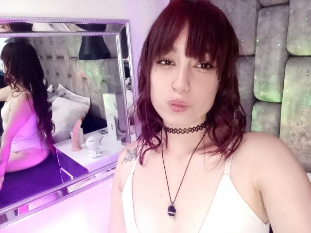 JennInk, com cabelo ruivo, camiseta branca, colar preto, piercing no nariz e colar, mostrando um reflexo de sua bunda em um espelho, com um brinquedo sexual rosa na cama..
